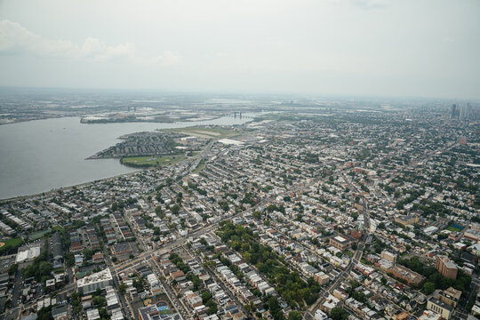 A high aerial view shows a dense residential neighborhood with streets and houses stretching across the city. Waterways and distant skyscrapers create a large metropolitan skyline on the horizon.