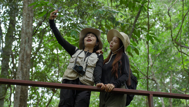 Two women hikers pose for a smartphone selfie beneath green forest trees, smiling together while documenting friendship and memorable moments during their outdoor nature travel adventure.