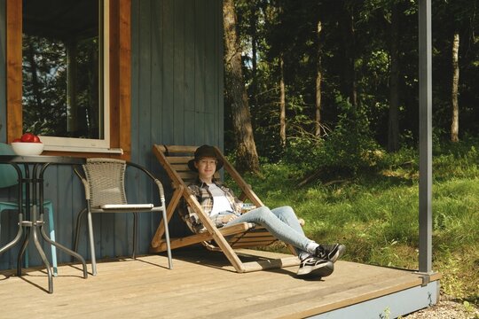 Cheerful young woman relaxes on deck chair outside wooden cabin in forest. Small table with fruit sits nearby
