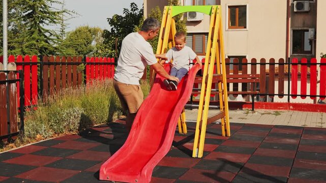 Happy father crouching down to catch his smiling toddler son sliding down a red slide on a playground on a sunny summer day.