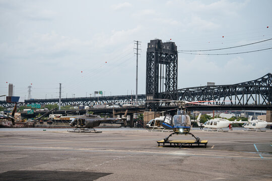 Several helicopters are parked on a large heliport near a river and steel bridge in an urban environment. The aviation area serves sightseeing flights and helicopter transportation.