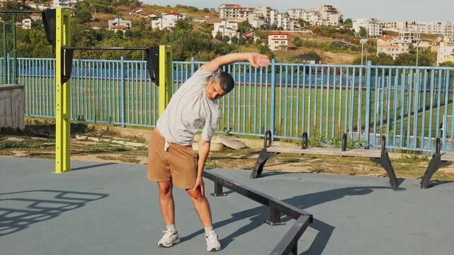 Adult man performing side bend body stretches at an outdoor sports park and gym facility on a bright sunny day.