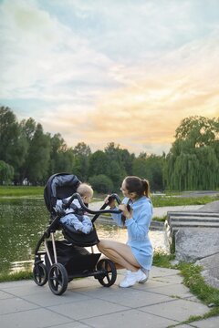 Mother is kneeling by stroller with her son on shore of calm water in urban park at sunset