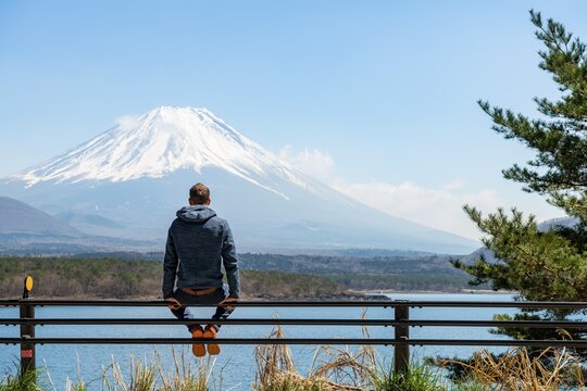 Young man sitting on railings next to a road and looking across the lake to Mt Fuji volcano, Motosu Lake, Yamanashi Prefecture, Japan