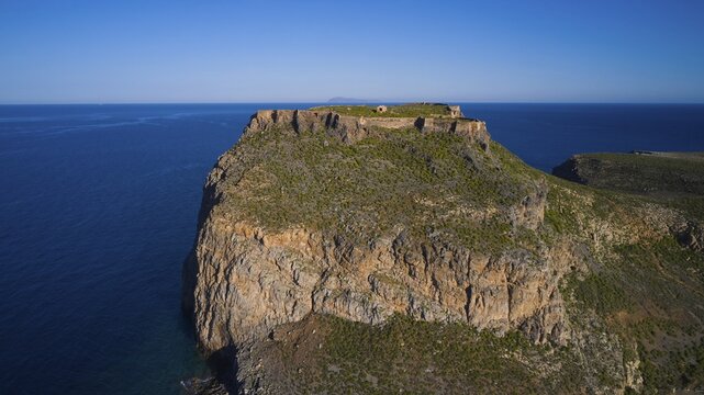 Drone shot, A dramatic, rocky coastline juts into the blue sea with lush vegetation on the cliffs, Venetian Sea Fortress, Gramvoussa, Gramvoussa Peninsula, Pirate Bay, Balos, Lagoon, Northwest Crete, Crete, Greek Islands, Greece