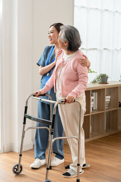 Young Asian nurse supports senior Asian woman standing with walker during rehabilitation, helping mobility, balance, safety and independence inside modern assisted living senior care center.