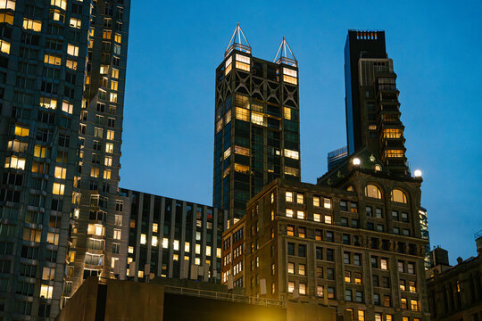 Tall Manhattan skyscrapers and historic buildings glow with warm lights against a deep blue evening sky. The illuminated architecture highlights the dramatic skyline of New York City at night.