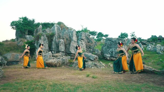 A wide shot showing a line of female dancers performing perfectly synchronized hand gestures and footwork in a beautiful limestone garden.