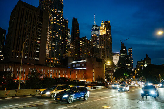 Cars drive along a busy Lower Manhattan roadway surrounded by illuminated skyscrapers at night. Bright headlights and city lights create a vibrant urban traffic scene.