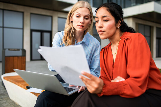 Two businesswomen reviewing documents outdoors with laptop