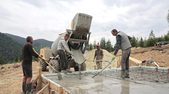 Four workers pour wet concrete from a mixer onto steel rebar flooring, The crew spreads fresh material inside timber forms for a new foundation