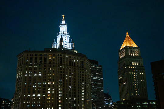 Historic towers and skyscrapers glow with warm lights against a deep blue night sky in Lower Manhattan. The illuminated skyline highlights the classic architecture of New York City.