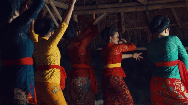 An angled rear shot of women in various colored Kebayas performing synchronized traditional Javanese dance moves inside a bamboo shelter.
