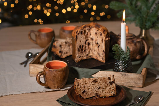 Table with sliced panettone bread pieces on plate, cups with tea, lighting candle and Christmas fir tree lights on background, traditional holiday food