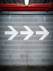 Fototapeta premium Three white arrow symbols painted on gray cinder block wall. Directional signs point right in industrial building underground parking lot. Concrete wall with red pipes above shows way to exit
