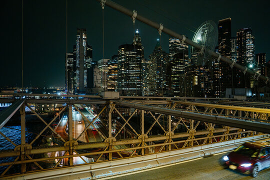 Cars drive across the Brooklyn Bridge while the illuminated Manhattan skyline rises in the background at night. Bright city lights and busy traffic create a dynamic urban scene in New York City.