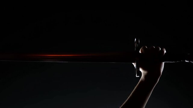 A hand grips a horizontally held sword as its blade moves through the semi-darkness