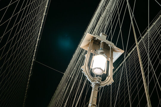 Historic street lamp glowing on Brooklyn Bridge surrounded by suspension cables against the dark night sky. Architectural details of the famous New York bridge with dramatic perspective and urban