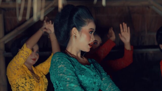 A side profile close-up of a female dancer in a green lace Kebaya and traditional bun, performing graceful arm movements with other dancers blurred.