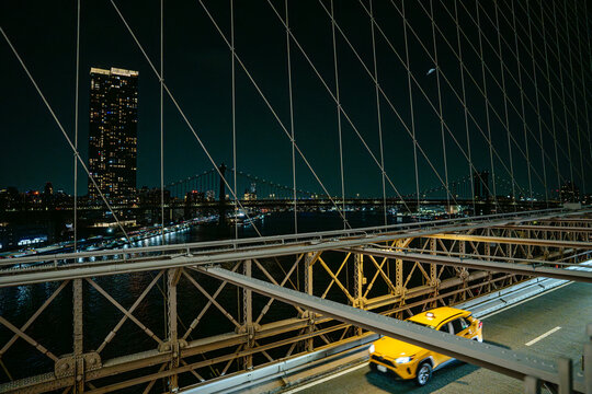 Yellow taxi driving across the Brooklyn Bridge with Manhattan skyline and illuminated buildings in New York City at night. Urban traffic scene with bridge cables, city lights, and reflections over the