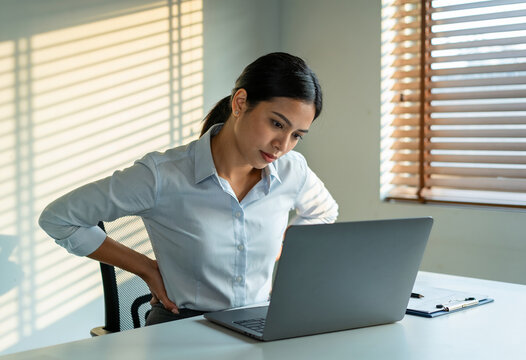 Asian businesswoman suffering lower back pain sitting at desk working on laptop in modern office natural sunlight