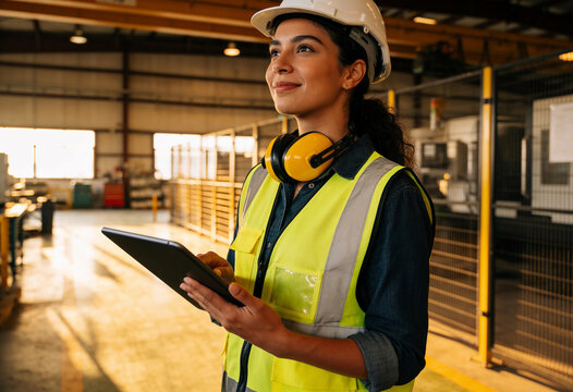 Confident female factory supervisor with safety gear holding digital tablet in industrial warehouse warm light