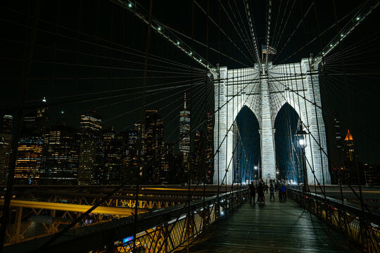 People walking along the Brooklyn Bridge pedestrian walkway with illuminated Manhattan skyline at night. Urban architecture scene with suspension cables and glowing city skyscrapers.