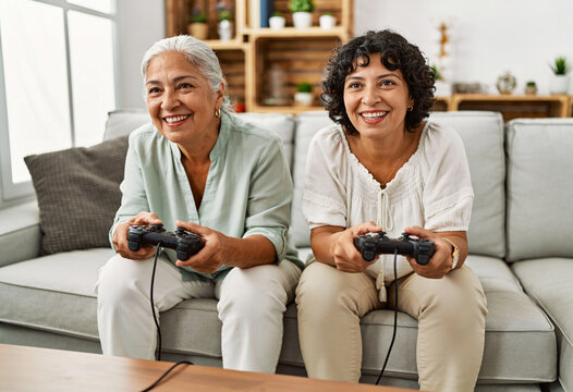 Senior Hispanic mother and adult daughter playing video games on sofa in bright living room holding controllers
