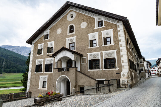 A grand traditional Engadine house in S-Chanf with ornate white sgraffito decoration on a cobblestone street with mountains behind in Switzerland