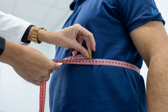 Doctor's hands measuring an adult man's waistline with a tape measure, checking his body dimensions for weight control and health