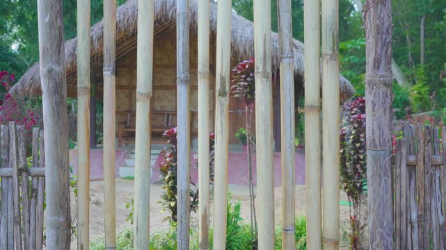Vertically arranged bamboo poles secured to horizontal beams form a village boundary, with a thatched dwelling and tropical vegetation beyond, illustrating locally sourced materials and rural resident