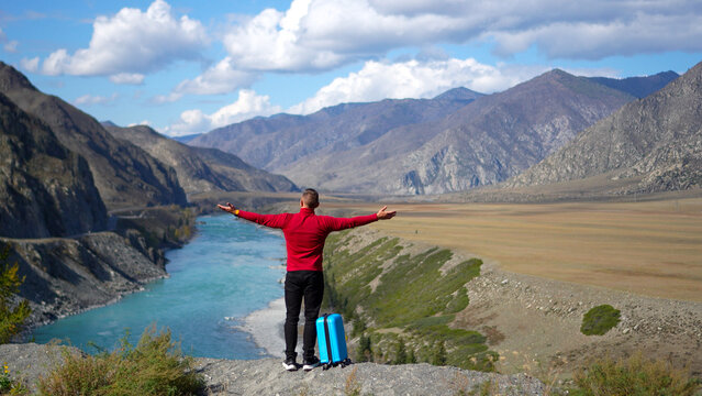 Exited traveler in red sweater raising hands with blue suitcase over river canyon.
