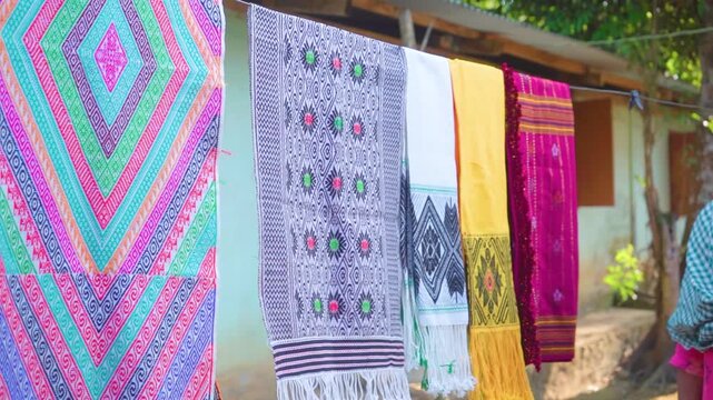 Colourful woven fabrics featuring geometric and floral designs hang outside a rural workspace, presenting finished textile products and the visual character of Assam's handloom sector in Shikdamakha, 