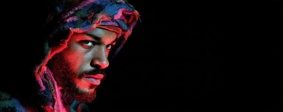 Man with intense gaze wearing colorful hooded garment in dark background, dramatic lighting highlights facial features and expression