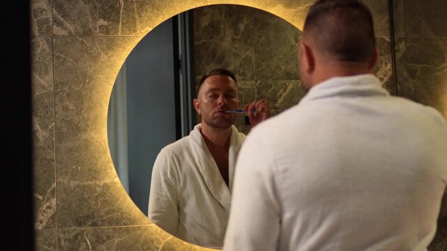Man in a white bathrobe checking his clean teeth looking in a round illuminated mirror with a static camera shot.