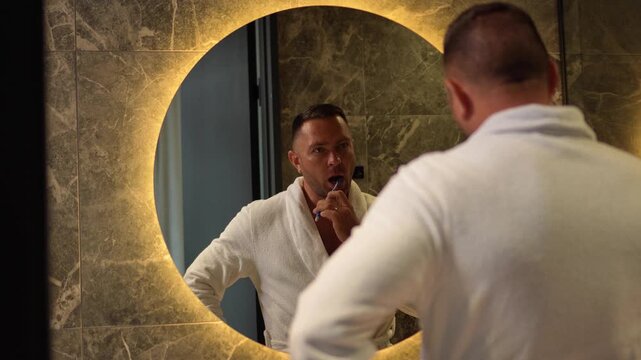 Man in a white bathrobe brushing his teeth looking in a round illuminated mirror with a static camera shot.