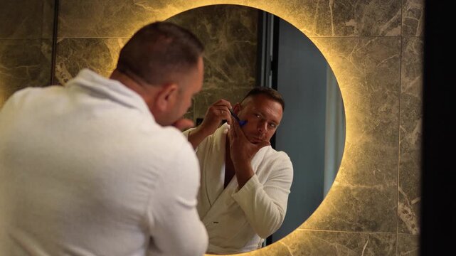 Handsome man in a white bathrobe shaving his face with a razor looking in a round illuminated mirror with a static camera shot.