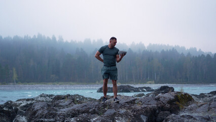 Fototapeta premium Front view of a strong young male tourist practicing strength workout and lifting a rock on a misty morning in Altai