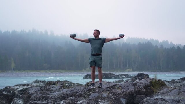 Front view of an athletic man standing on river rocks with arms outstretched holding stones against a foggy forest
