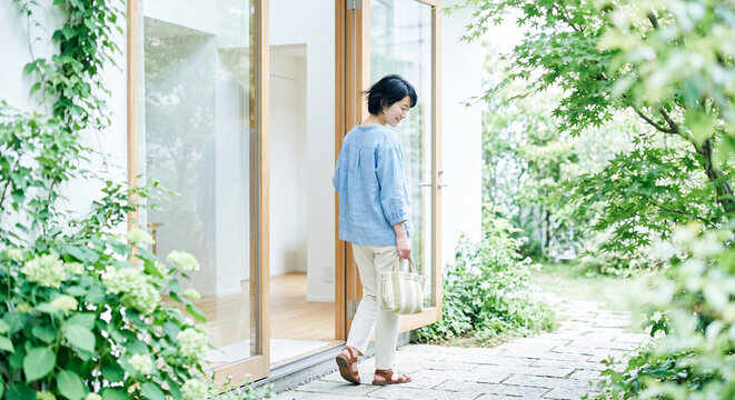 Woman strolling through vibrant green foliage and vines toward a modern glass house entrance in dappled sunlight. Fresh architecture and lifestyle concept.