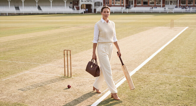 Woman in a classic white cricket uniform and cap walking across a green cricket pitch carrying a bat and bag. Elegant sport and lifestyle fashion concept.
