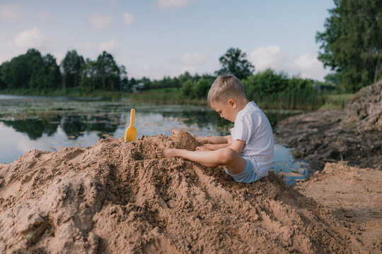 Young boy playing with sand near a calm lake, wearing a white t-shirt and blue shorts, with a yellow plastic shovel beside him on a sunny day