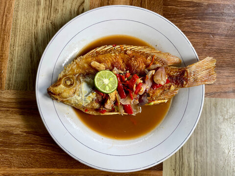 Fried *mujair* fish prepared as "Pecak Ikan," topped with a traditional spiced broth and lime wedges, served on a white plate atop a wood-patterned table.