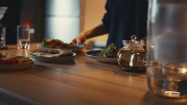 Woman serving healthy dinner for two on a wooden kitchen table, placing two plates with fresh salad. Glass teapot with brewing herbal tea, open cheese sandwiches and sliced fruits in a warm cozy home.