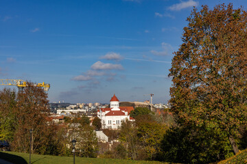 Fototapeta premium Panoramic autumn view of Vilnius, Lithuania, on a sunny October day, featuring a historic white church with red tiled roofs surrounded by colorful autumn trees, with construction cranes.