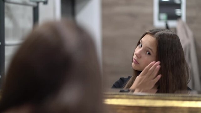Young brunette girl combs hair in front of mirror, daily morning beauty routine and hair care at home.