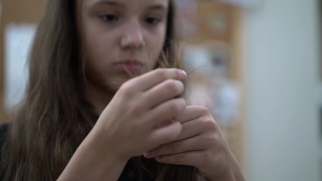 Teenage girl examines split ends of her long brown hair held in both hands, slight indoor smile.