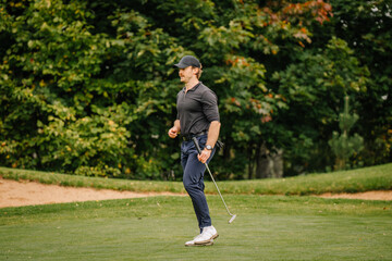 Obraz premium Golfer walking across manicured grass holding a club beside a sandy path and lush green trees in an outdoor recreational landscape scene