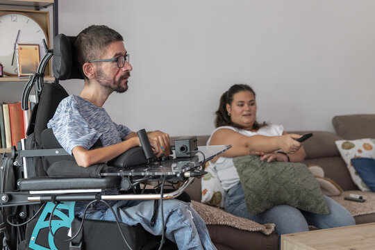 A man in a wheelchair sits in a living room while a woman assists him with watching TV. They are in a comfortable home setting.
