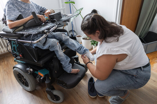 A woman helps a man in a wheelchair with putting on his socks and shoes at home. They are in a well-lit room with plants.